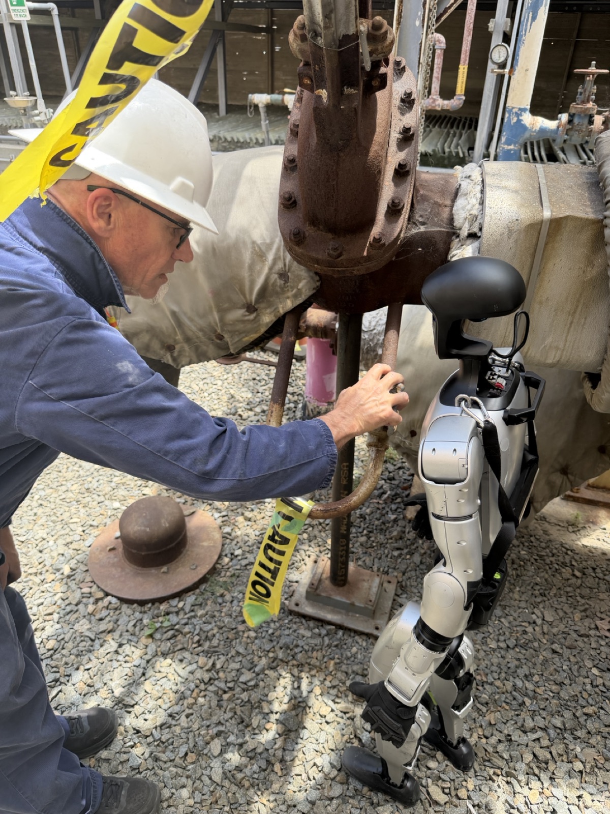 An operator adjusting a valve by hand while a humanoid robot works alongside at a power plant